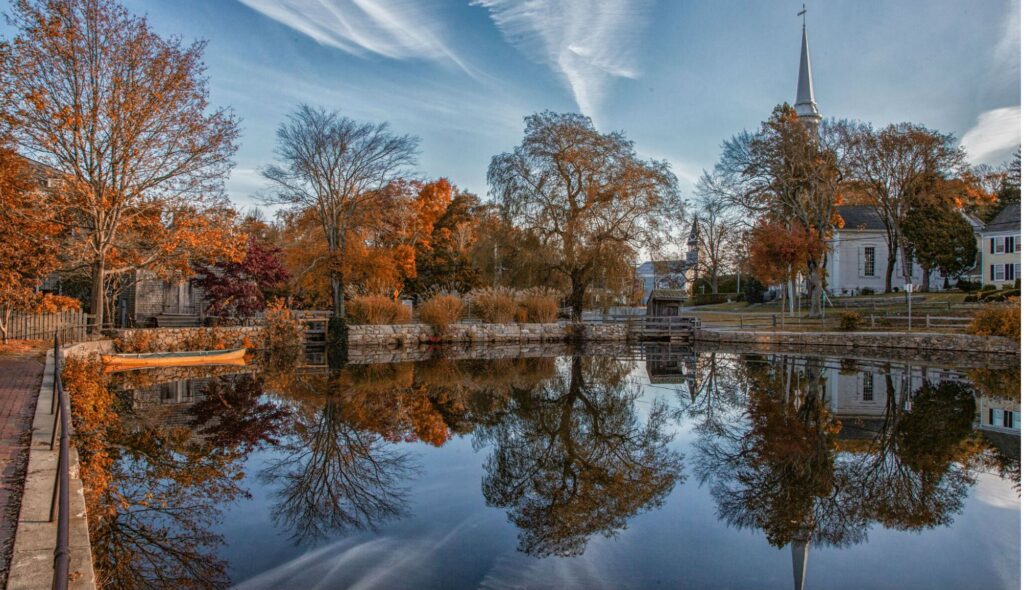 An image of downtown Sandwich, Massachusetts. It showcases the fall foliage and pond. 
